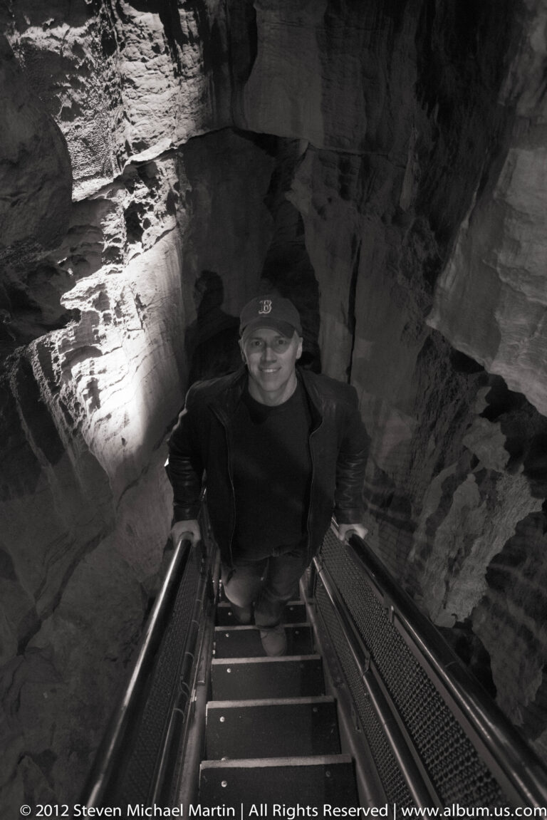 A visitor climbs a narrow metal stairway between close cave walls, holding the railings.