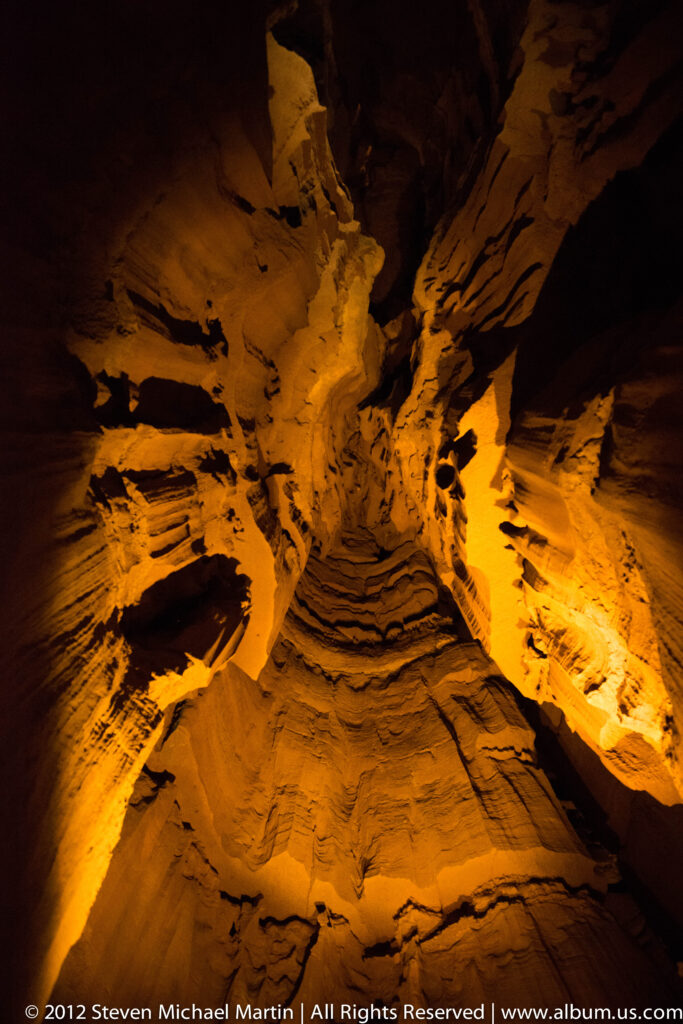 Amber light reveals ridged cave walls and hanging formations around a dark opening above.