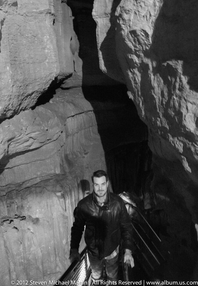 A man pauses on a metal stairway in a cave, framed by bright rock and a dark recess.
