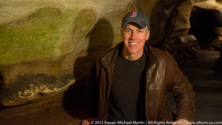 A smiling visitor in a dark cap and brown jacket stands before a dimly lit cave wall.
