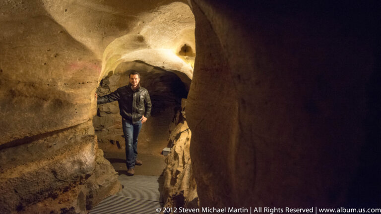 A visitor stands on a metal walkway in a narrow corridor between curving cave walls.