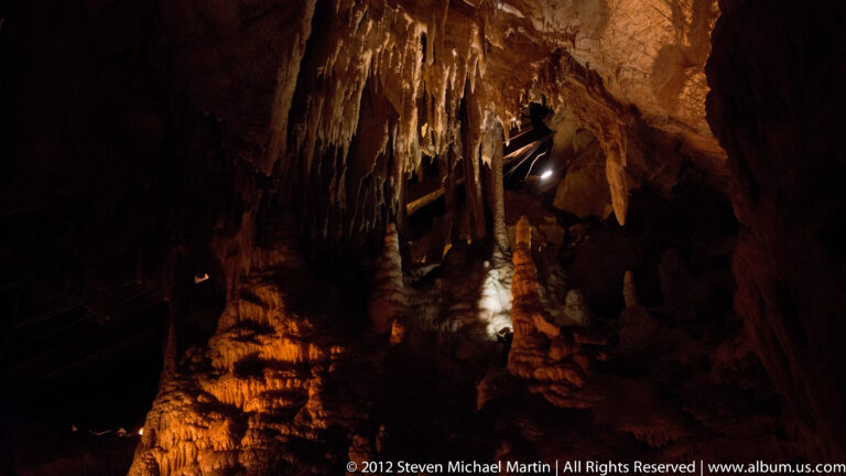Long stalactites descend toward a shadowed floor, with a faint visitor rail at left.