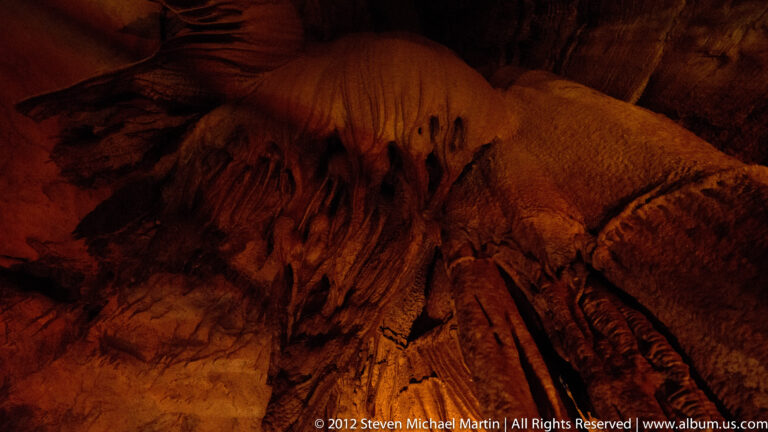 A rounded mineral mound with parallel ridges glows red-orange beneath small hanging formations.