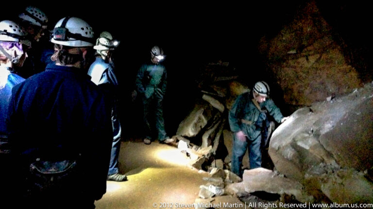 Helmeted visitors with headlamps gather around a boulder in a dim cave passage.