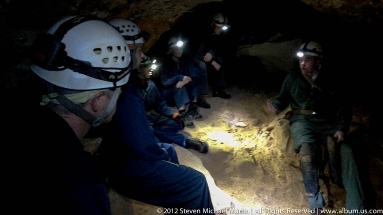 Helmeted visitors sit along a low cave wall while headlamps illuminate the uneven floor.