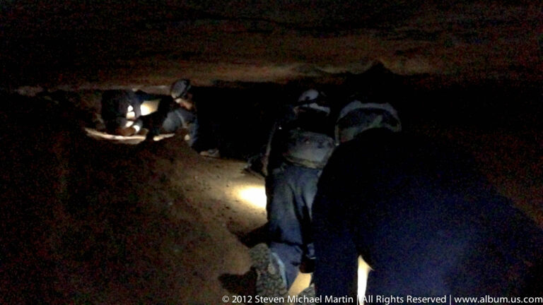 Helmeted visitors crouch beneath a low rock ceiling and move toward a lit opening.