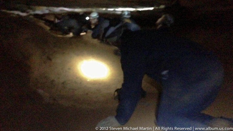 Visitors crouch beneath a low cave ceiling as a headlamp beam lights the floor ahead.