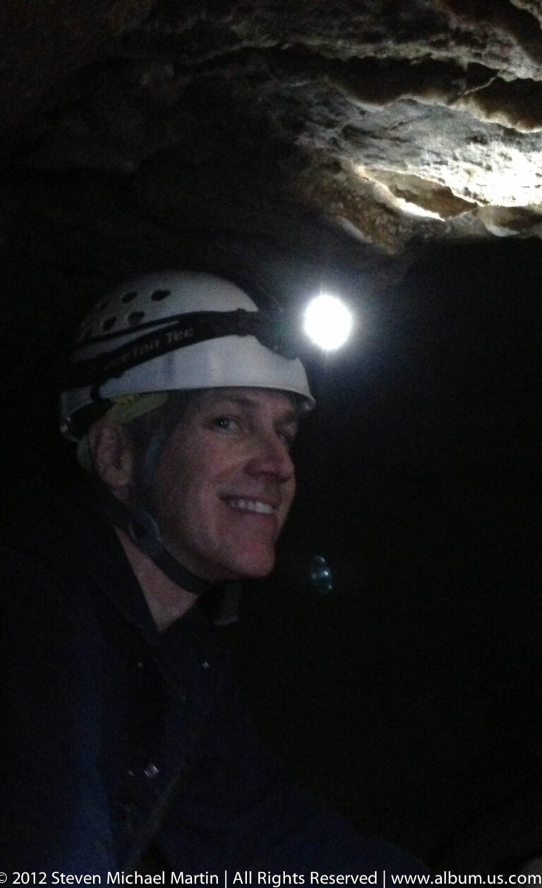 A smiling visitor in a white helmet and headlamp poses against a dark cave interior.