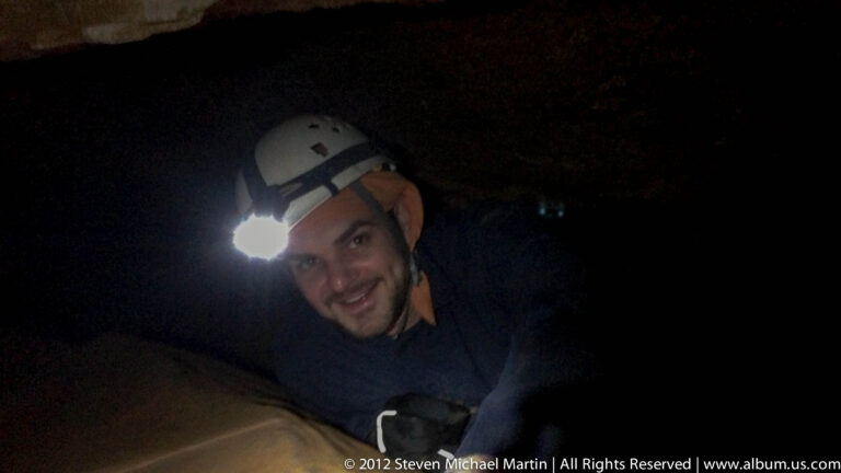 A smiling man wearing a white helmet and headlamp leans close to the camera in a dark cave.