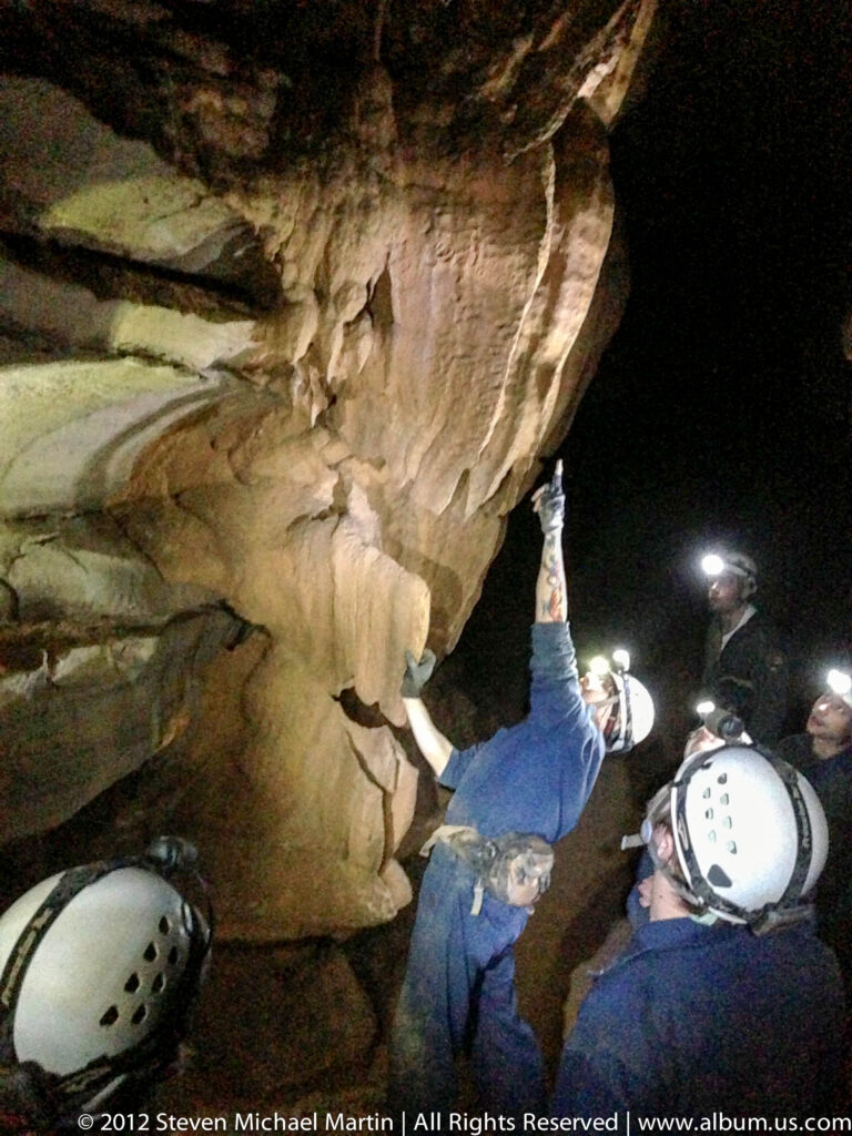 Helmeted visitors cluster beneath a low limestone ceiling; one raises an arm toward the rock.