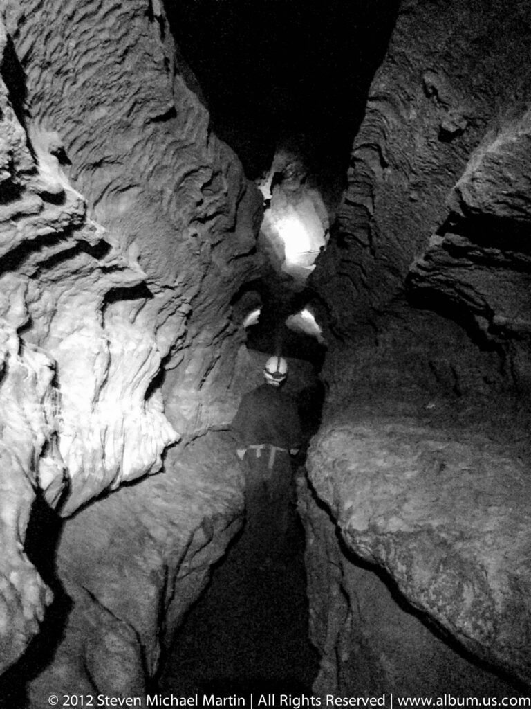 A dark cave chamber is framed by layered rock, with a bright patch of light on the far wall.
