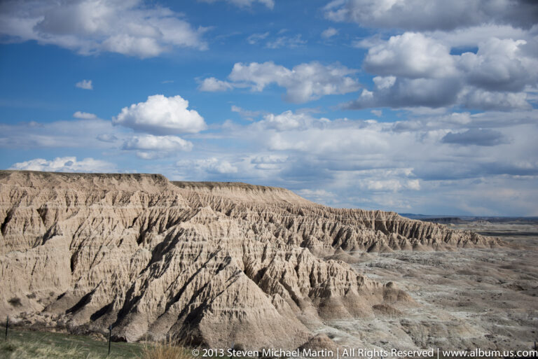 Pale eroded ridges and branching gullies stretch beneath large white clouds across a blue sky.