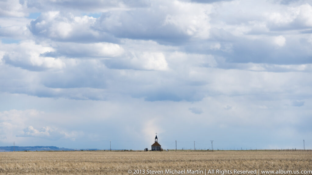 A small white church with a steeple stands on flat prairie beneath a broad cloudy sky.