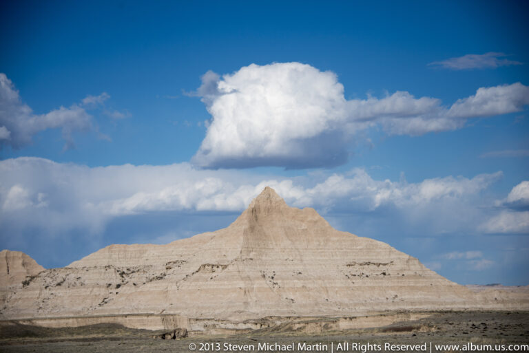 A pale steep-sided butte stands beneath a towering white cloud in a deep blue sky.