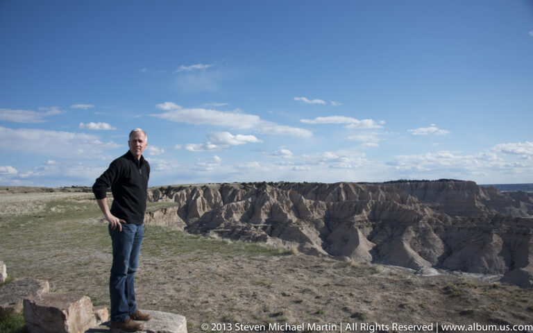 A man stands on a rock ledge above steep badlands ridges beneath a broad blue sky.