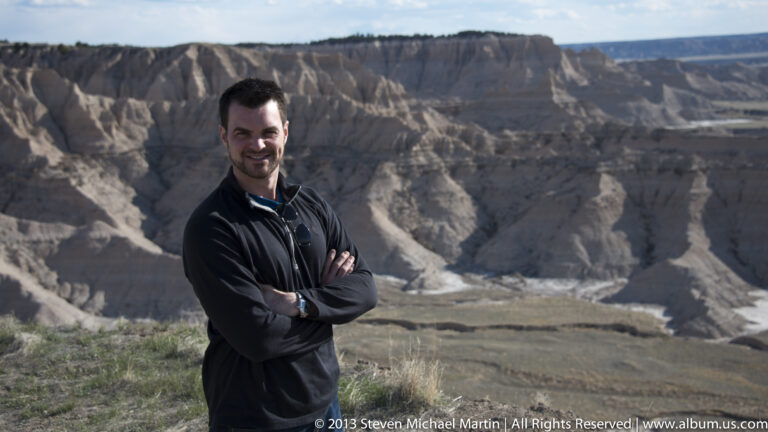 A man in a dark jacket poses with folded arms before pale eroded badlands ridges.
