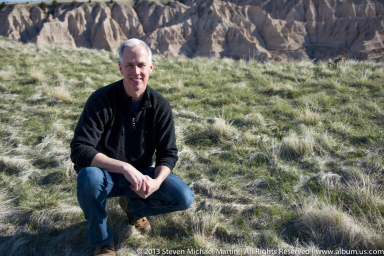 A crouching man smiles on a grassy rim with steep eroded ridges behind him.