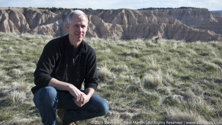 A seated man rests on a grassy slope with jagged pale badlands ridges behind him.