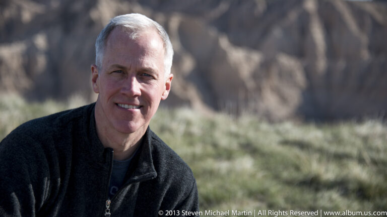 A close portrait of a man looking at the camera, with softly blurred badlands behind him.