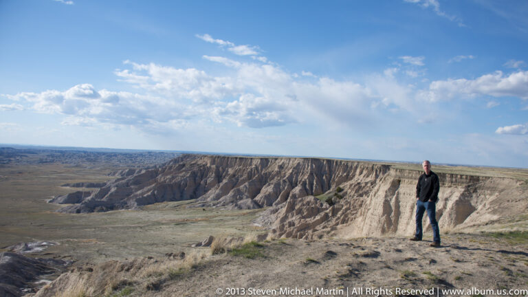 A man stands at the edge of a broad overlook above a long line of badlands ridges.