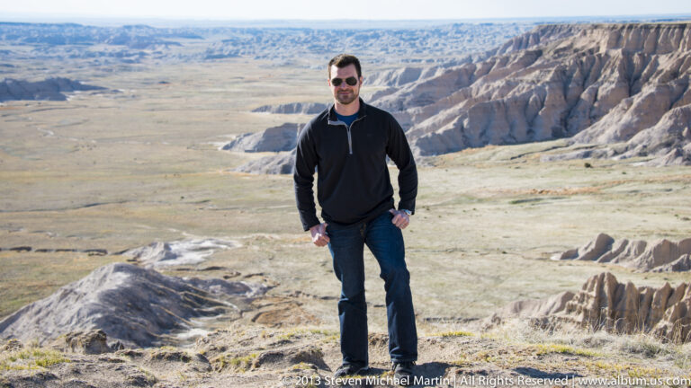 A man in sunglasses stands beside a boulder above a broad badlands basin.