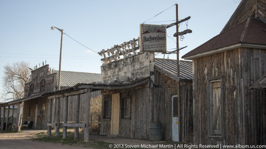 Weathered wooden storefronts carry faded painted lettering and a raised Budweiser sign above the roofs.
