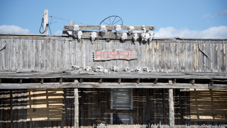 A faded red Hostel sign hangs on a gray timber building beneath mounted skulls and horns.