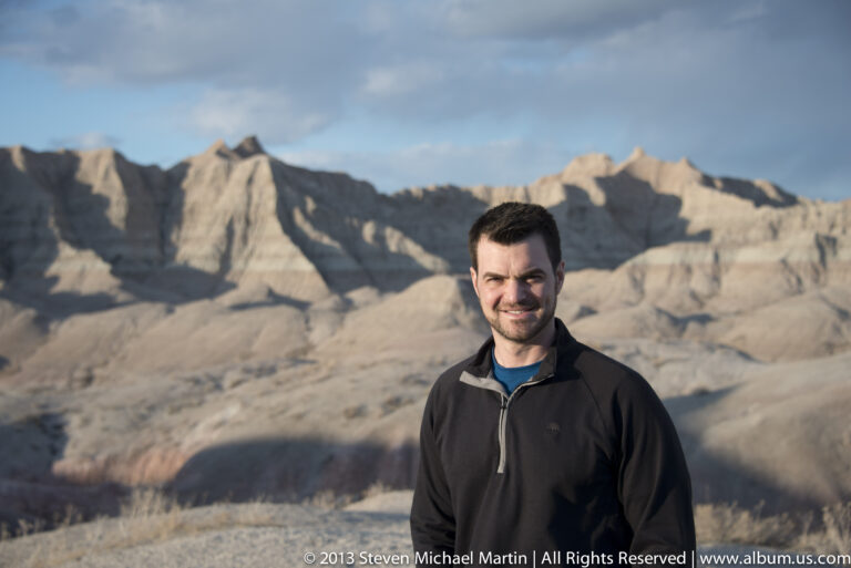 A man in a dark jacket poses before sunlit, sharply folded badlands ridges.