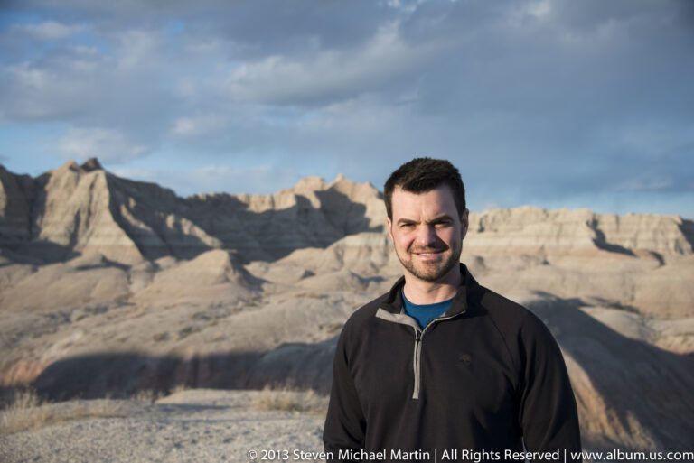 A close portrait of a man in a dark jacket and blue shirt against softly focused sunlit ridges.
