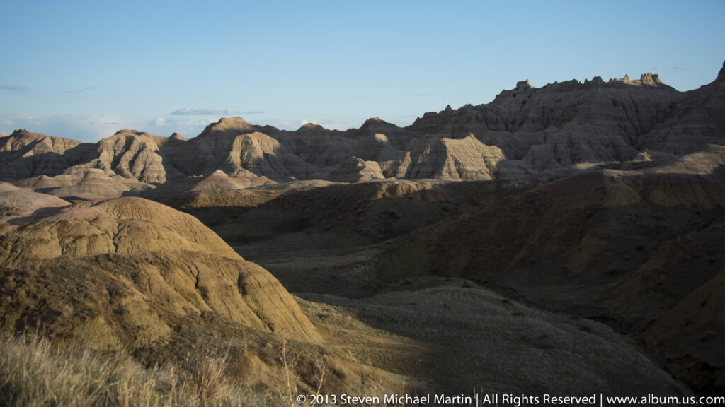 Shadowed badlands mounds lead toward sunlit ridges along the horizon at dusk.