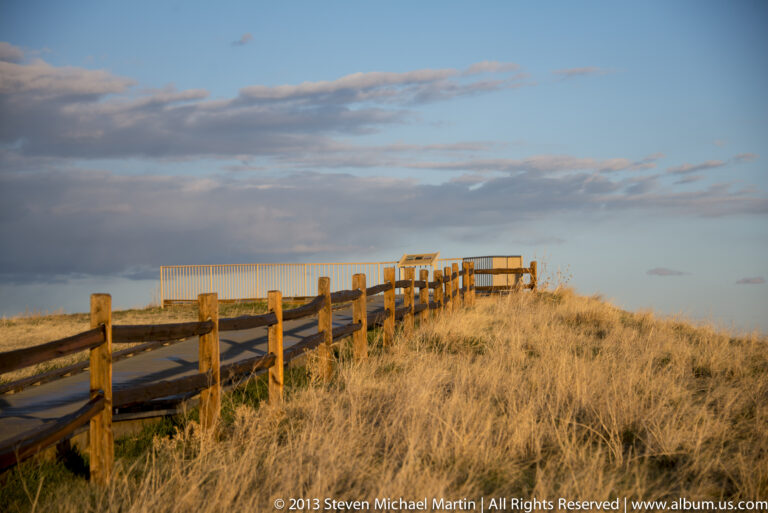 A wooden boardwalk crosses tall dry prairie grass toward a small platform beneath a cloudy sky.