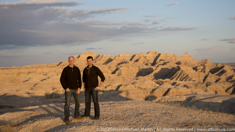 Two men pose together on gravel before sunlit jagged badlands formations in warm evening light.