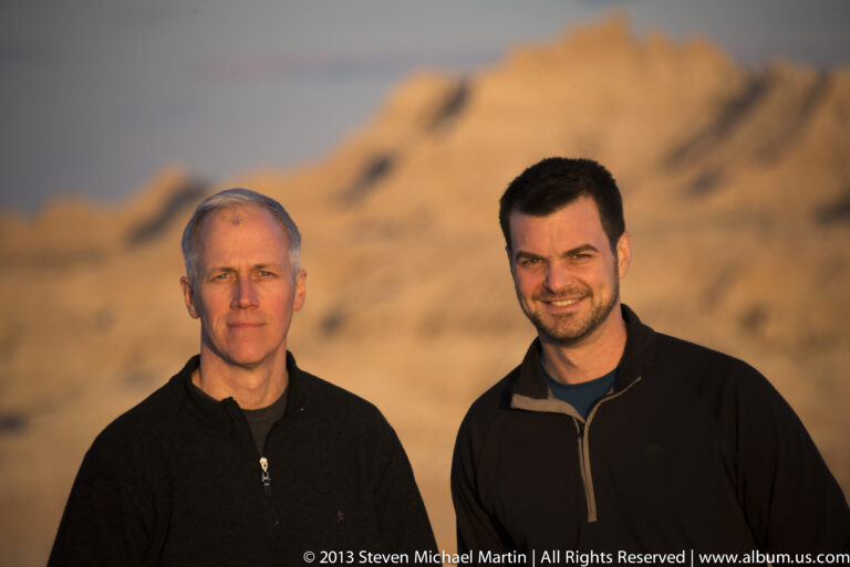 A close portrait shows two men against a softly blurred orange badlands formation at sunset.