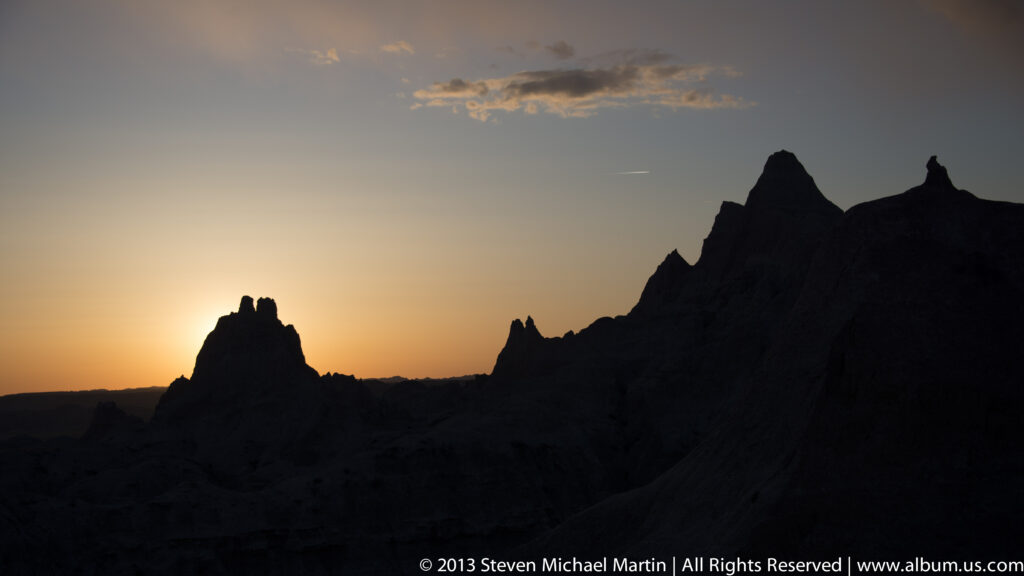 Dark jagged pinnacles silhouette against an orange and blue sunset sky with the sun near the horizon.