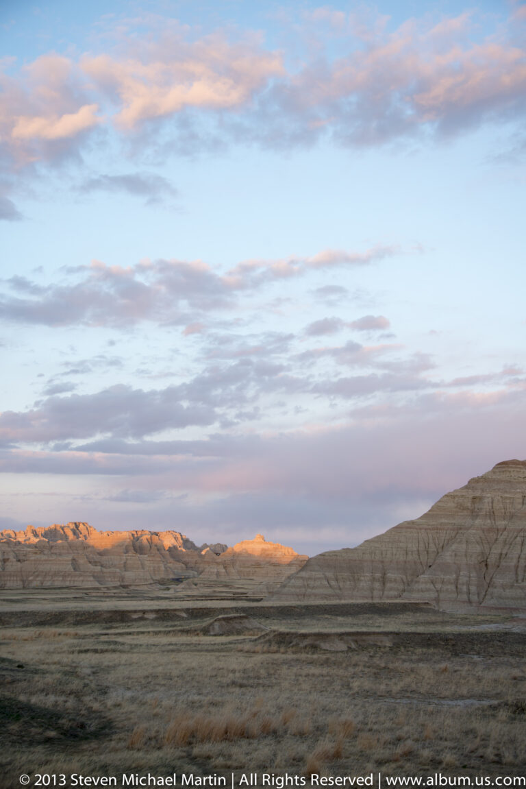 A dark prairie extends toward distant badlands ridges glowing in evening light beneath pink and blue clouds.