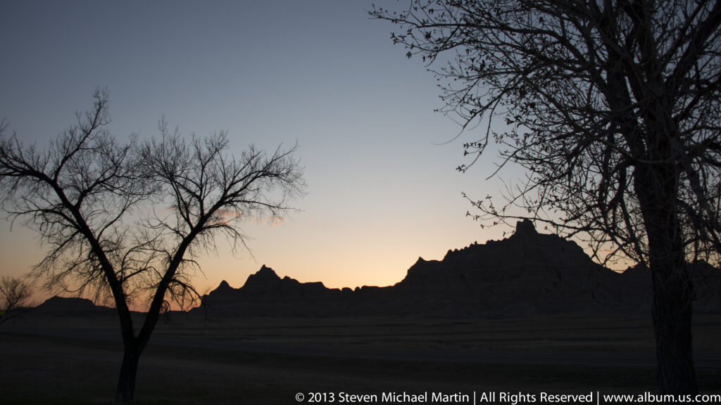 Leafless trees and a jagged badlands ridge form dark silhouettes against a peach sunset horizon.