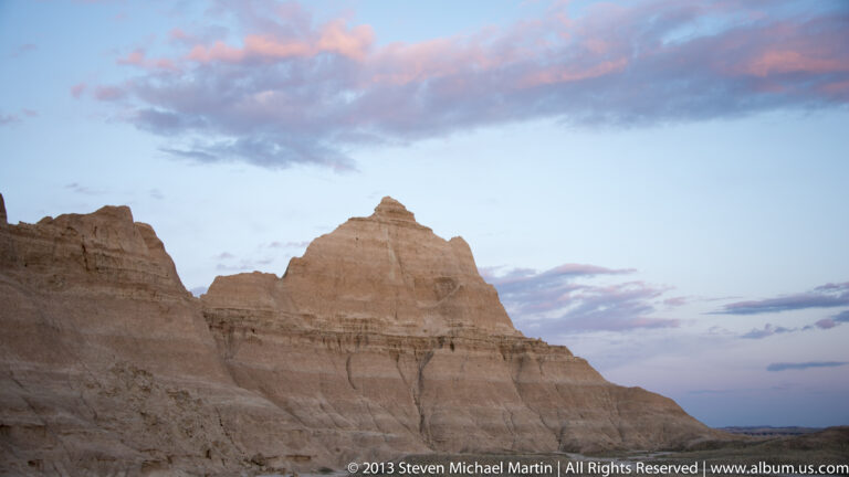 A banded butte rises between eroded rock walls beneath a blue sky streaked with pink clouds.