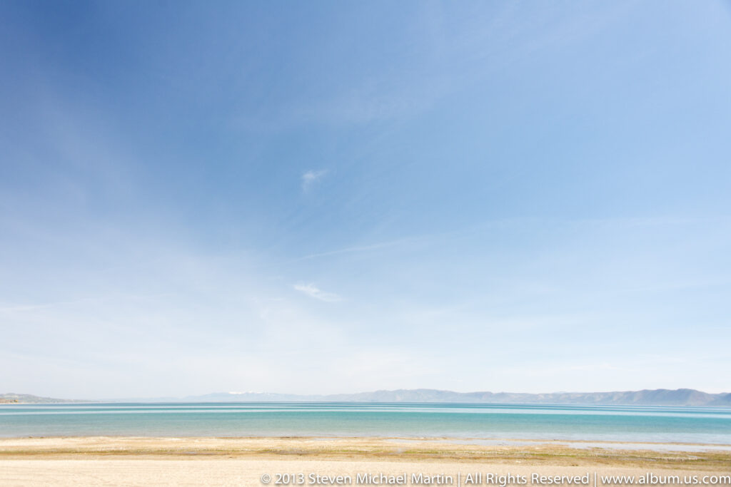 Turquoise water and a pale sandy shore extend toward hazy low mountains beneath a broad blue sky.