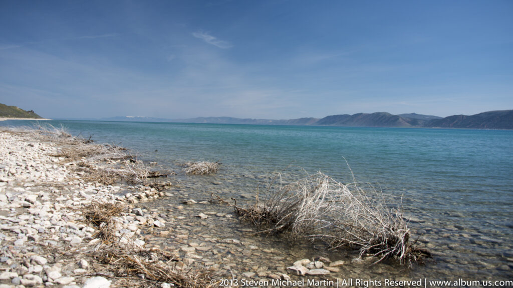 Clear water laps over pale stones along a dry, brush-lined shore, with distant mountains across the lake.