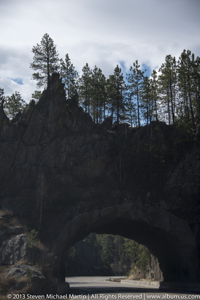 A dark natural rock arch crosses a forested ridge, with pine silhouettes along the bright skyline.