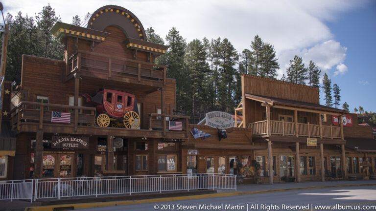 Wooden storefronts include a red stagecoach above a balcony, flags, and theater outlet signs.
