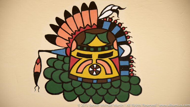 A colorful outlined figure with a feathered headdress and geometric face is painted on a pale background.