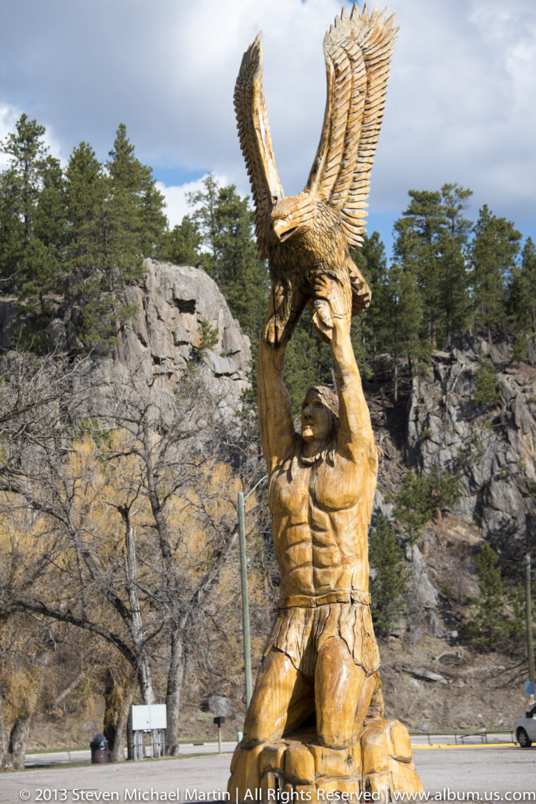 A carved wooden person lifts a spread-winged eagle above their head outdoors among pines.