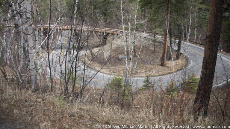 A paved road forms a sharp hairpin turn around a wooded slope beneath another road.