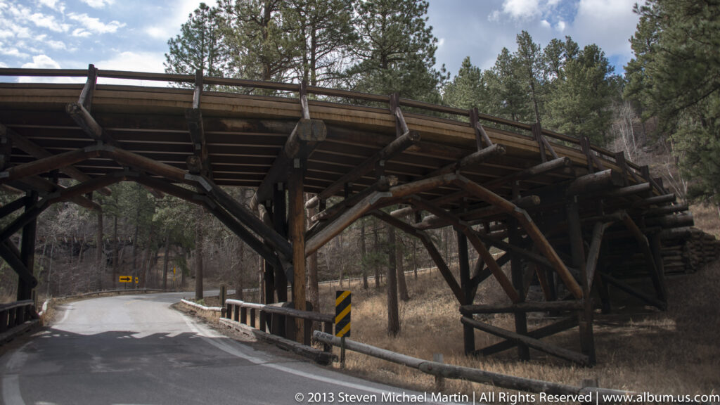 A timber trestle with exposed braces spans above a curving paved road in a wooded ravine.