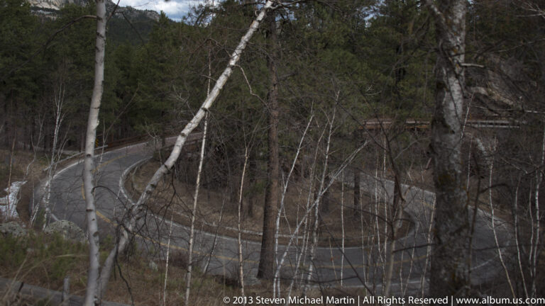 A winding paved road doubles back across a wooded hillside, partly hidden by bare trees.