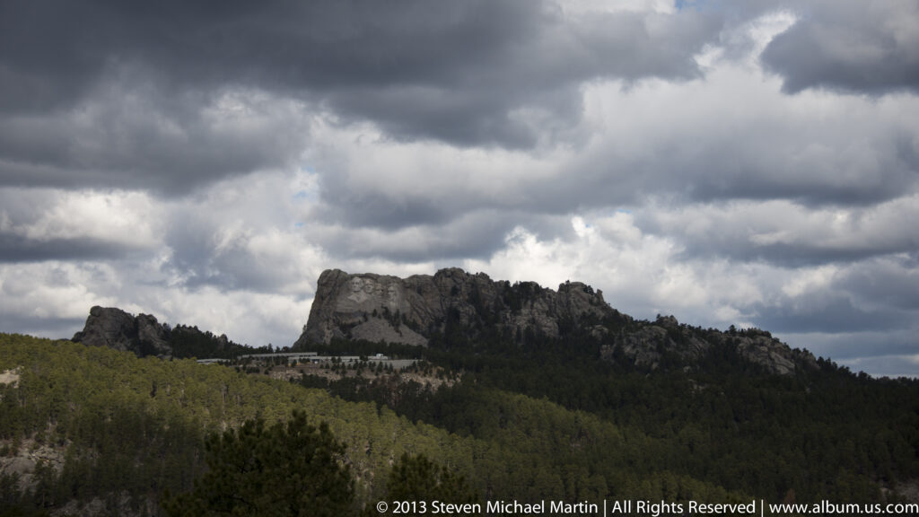 Mount Rushmore appears along a forested granite ridge beneath a broad bank of storm clouds.