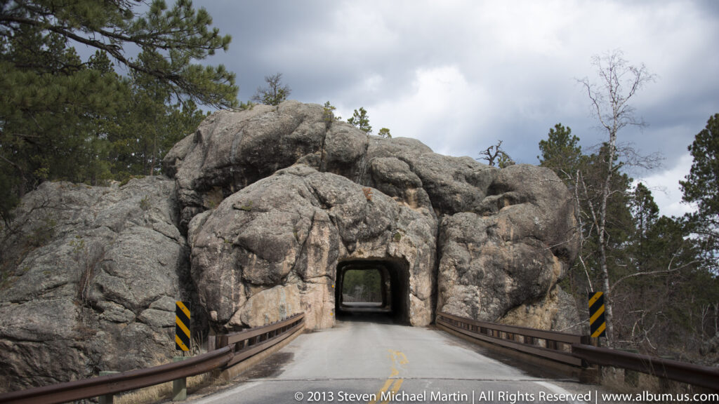 A paved road enters a rectangular tunnel cut through a granite outcrop, flanked by guardrails.