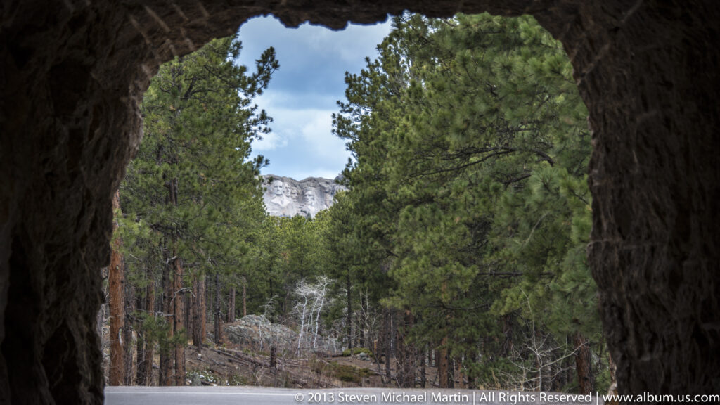 Pine trees and a pale rock face appear through a dark, irregular opening in the foreground.