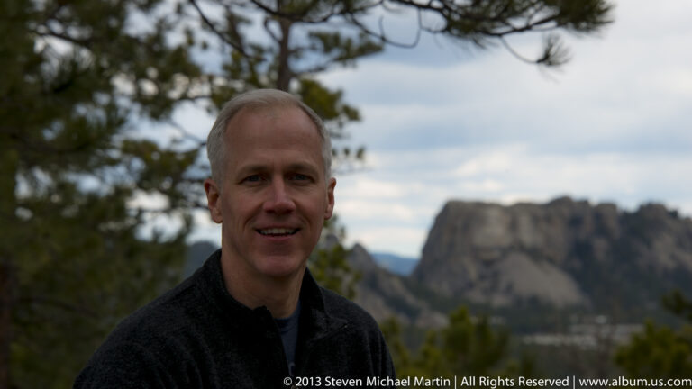 A man poses beneath pine branches with Mount Rushmore softly visible behind him.